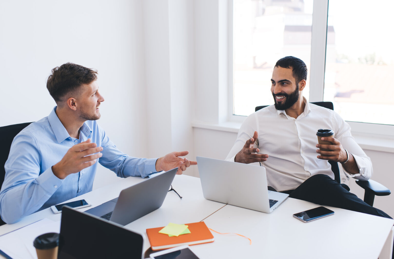 Cheerful male entrepreneurs in smart casual shirts smiling during collaborative working process in office interior with laptop computers on desktop, happy diverse colleagues talking about business